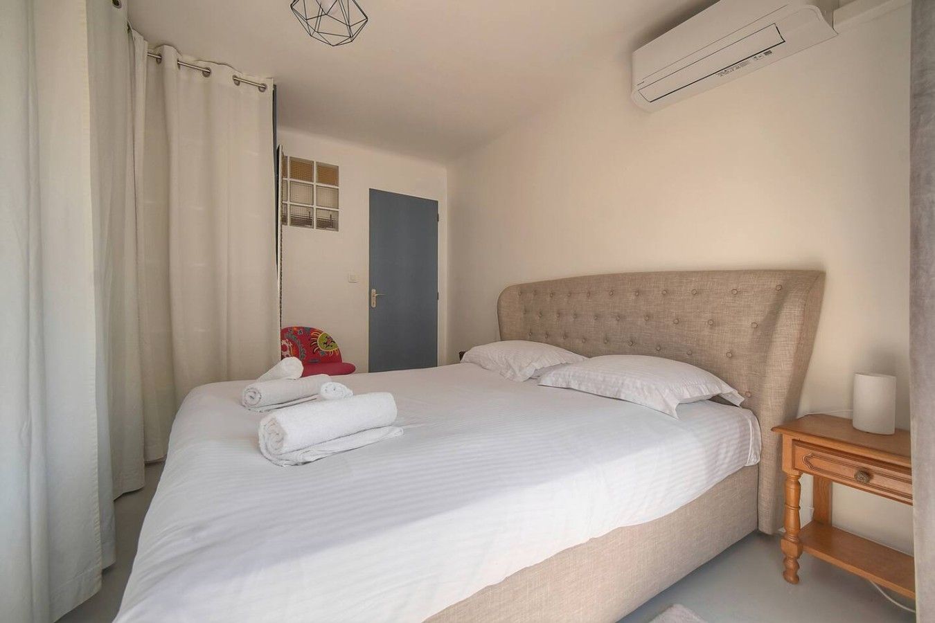 Une chambre à coucher comprenant un lit, des oreillers, une table de chevet, un appareil de climatisation, une porte et une fenêtre.