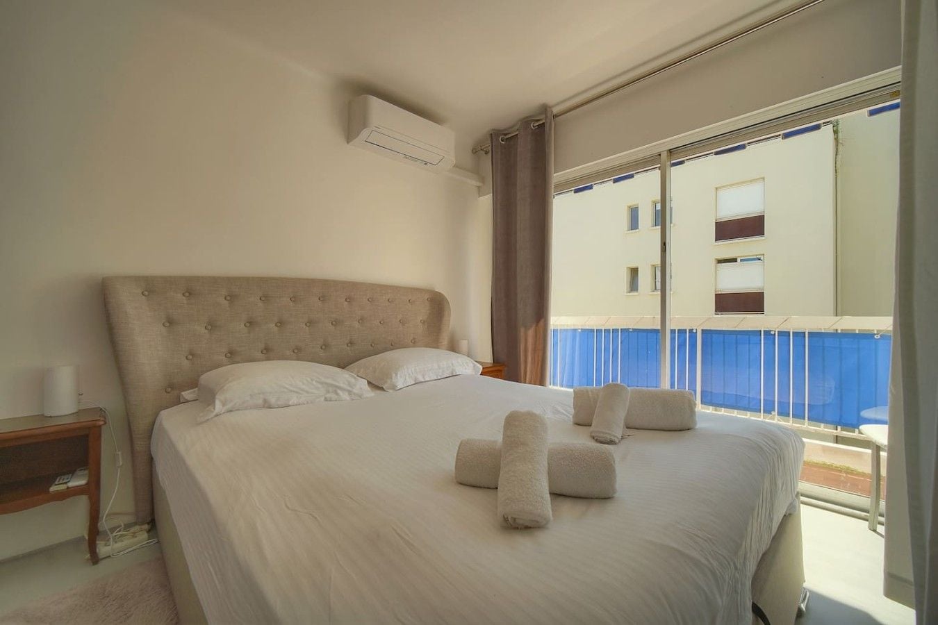 A hotel room with a large bed, a bedside table, an air conditioning unit, and a view of a building with blue balconies.