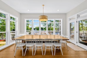 Un comedor espacioso con una gran mesa de madera rodeada de sillas blancas, ventanas amplias y una lámpara colgante.
