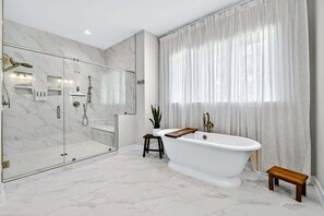 Ein modernes Badezimmer mit einer Glaskabine zur Dusche, einer freistehenden Badewanne und einem großen Fenster mit durchsichtigen Vorhängen.