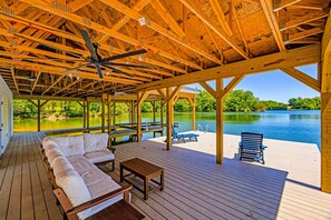 Una terraza techada al aire libre con techo de madera, un sofá blanco, una mesita y vista a un lago.