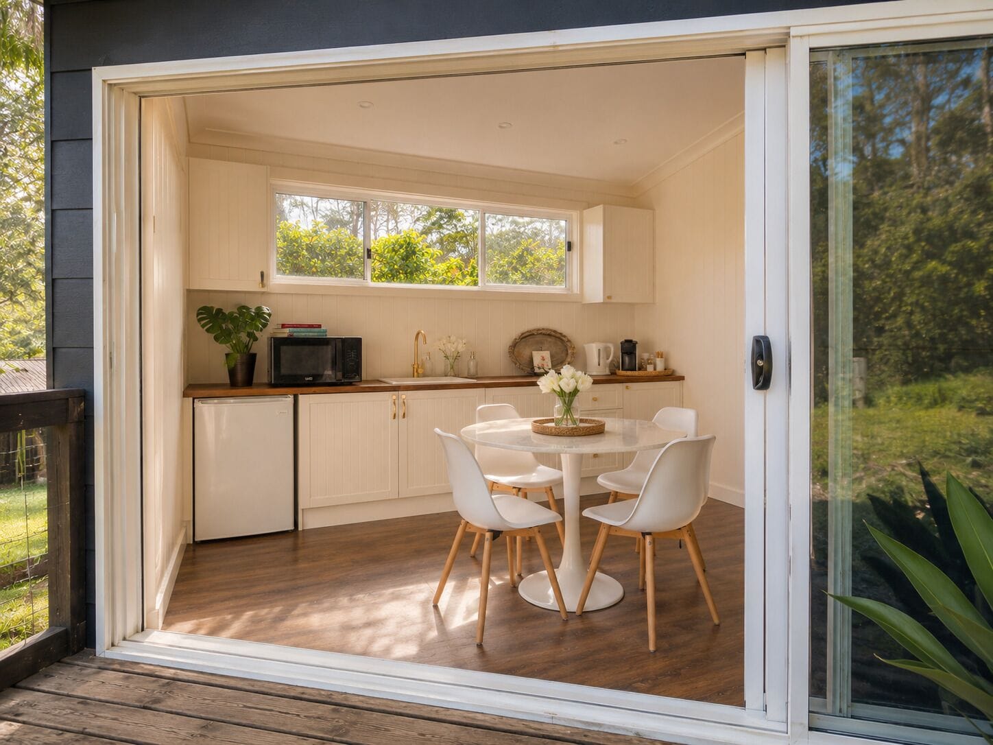 A modern kitchen with white cabinets, a dining table with white chairs, and a view of greenery outside.