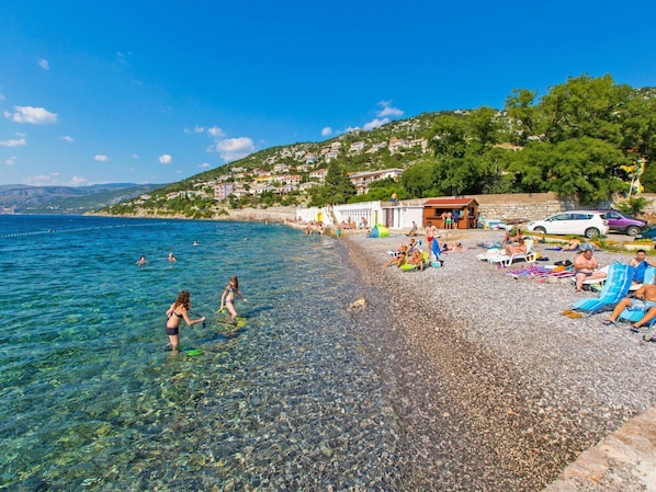 Ein Kiesstrand mit klarem Wasser, darin badende Menschen und Liegestühle.