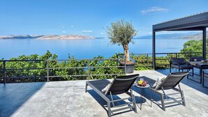 Een terras met twee comfortabele stoelen, een tafel met fruit en uitzicht op zee en bergen.