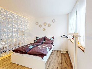 A bedroom with a bed, a white partition, a wall-mounted mirror, and a window with curtains. - Renovierte Erdgeschosswohnung by Interhome (Simmersfeld)