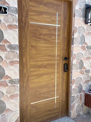 Uma porta de madeira com uma linha vertical e uma linha horizontal, uma parede de pedra e uma pequena prateleira com um aparelho de luz.