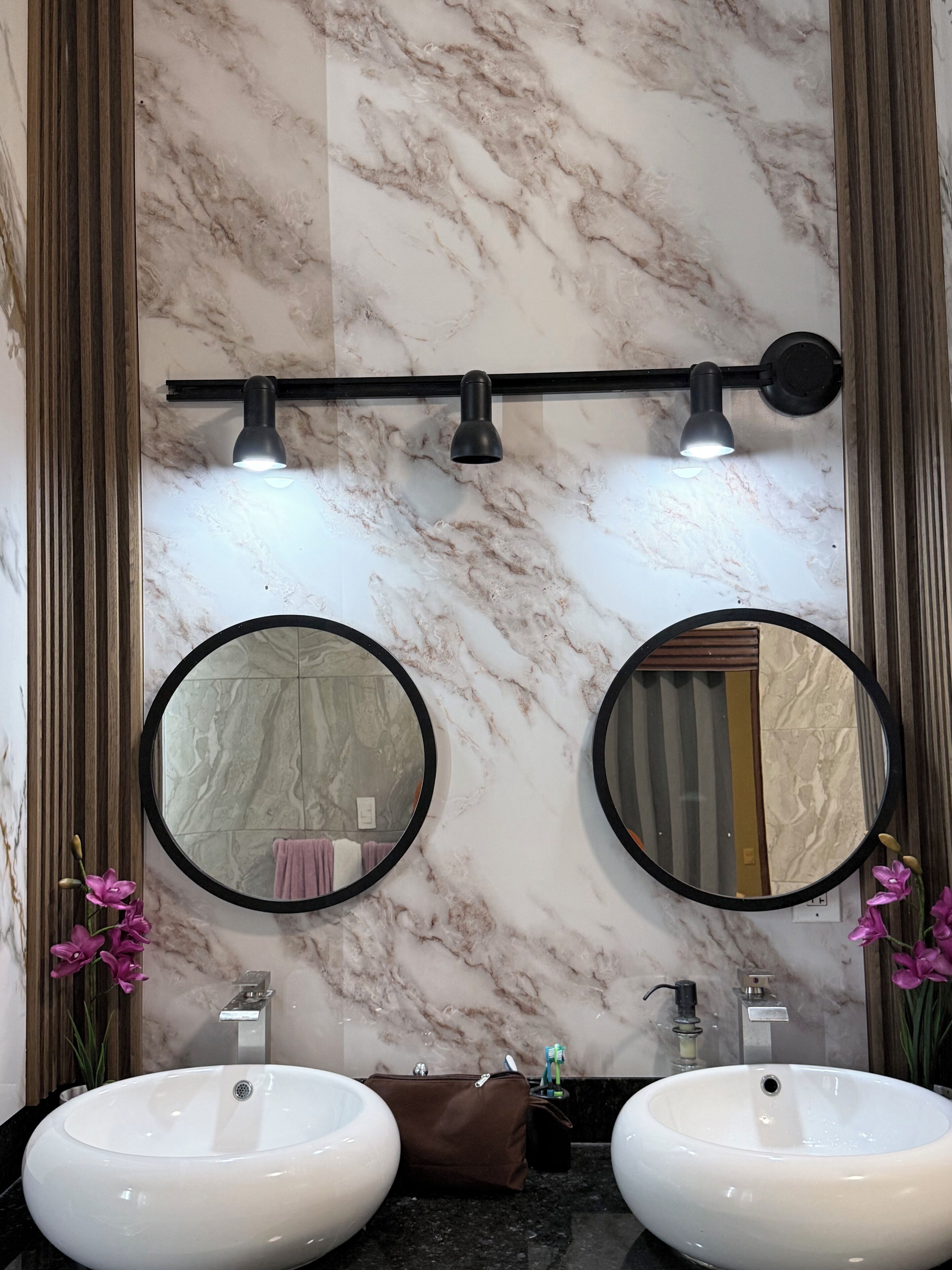 A bathroom with two round mirrors, a marble wall, a towel rack, and a sink with a brown bag on the counter.