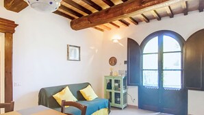 Ein Zimmer mit Holzbalkendecke, einem grünen Sofa mit gelben Kissen, einem Esstisch mit Stühlen, einem Schrank und einem großen Bogenfenster.