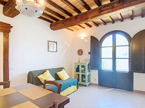 Ein Zimmer mit Holzbalkendecke, einem grünen Sofa mit gelben Kissen, einem Esstisch mit Stühlen, einem Schrank und einem großen Bogenfenster.
