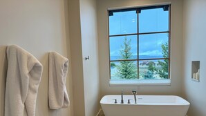 Un baño con una bañera independiente, un banco de madera y un ventanal con vistas a árboles y edificios.