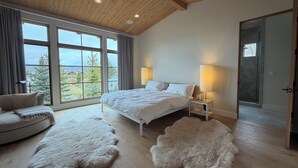 Un dormitorio con una cama grande, dos lámparas de noche, una alfombra mullida, un sofá y vistas a árboles a través de amplios ventanales.