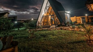 Un bâtiment moderne de forme triangulaire, doté d’une façade en verre, entouré d’une zone verdoyante et d’une structure au toit de chaume.