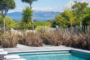 Una piscina con muelle de madera, rodeada de plantas y árboles, con un paisaje montañoso al fondo.