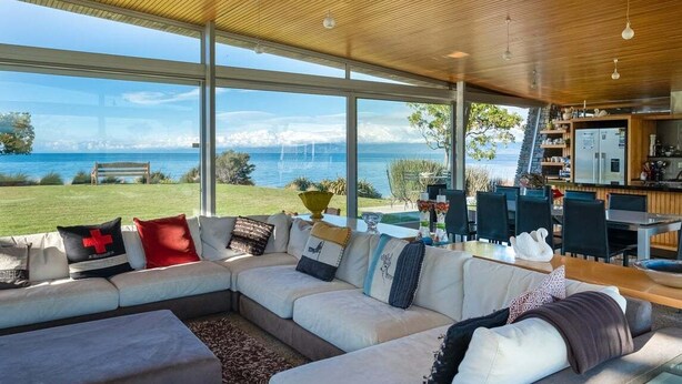 A modern living room with a large sectional sofa, a dining area, and a view of the sea. - Kina Retreat - Main House with Pool Access (Kina Peninsula)