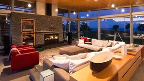 A modern living room with a stone fireplace, large windows, and a wooden ceiling. - Kina Retreat - Main House with Pool Access (Kina Peninsula)
