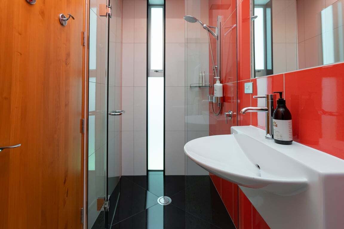 A modern bathroom with a glass shower enclosure, a white sink, and a red-tiled wall.
