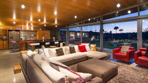 A modern living room with a large sectional sofa, red chairs, a kitchen area, and large windows offering a view of the outdoors. - Kina Retreat - Main House with Pool Access (Kina Peninsula)