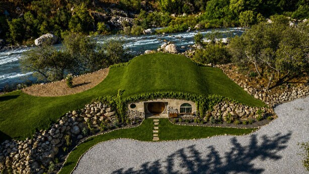 Uma casa com um telhado curvo peculiar, rodeada por um gramado bem cuidado e um rio.