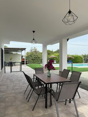 An outdoor patio with a table and chairs, a hanging light fixture, and a view of a swimming pool.