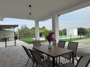 An outdoor patio with a table and chairs, a pool in the background, and a covered area with a grill.