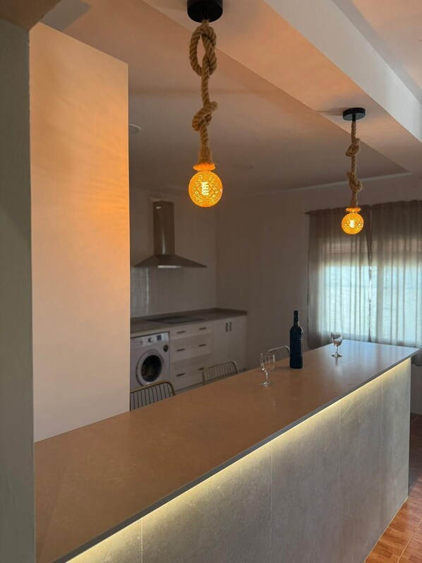 A modern kitchen with a central island, pendant lights, a washing machine, and a dining area with a wine glass and bottle.