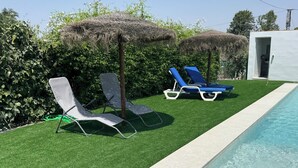 Zona de piscina con césped artificial, dos sillas de descanso y una sombrilla de paja.