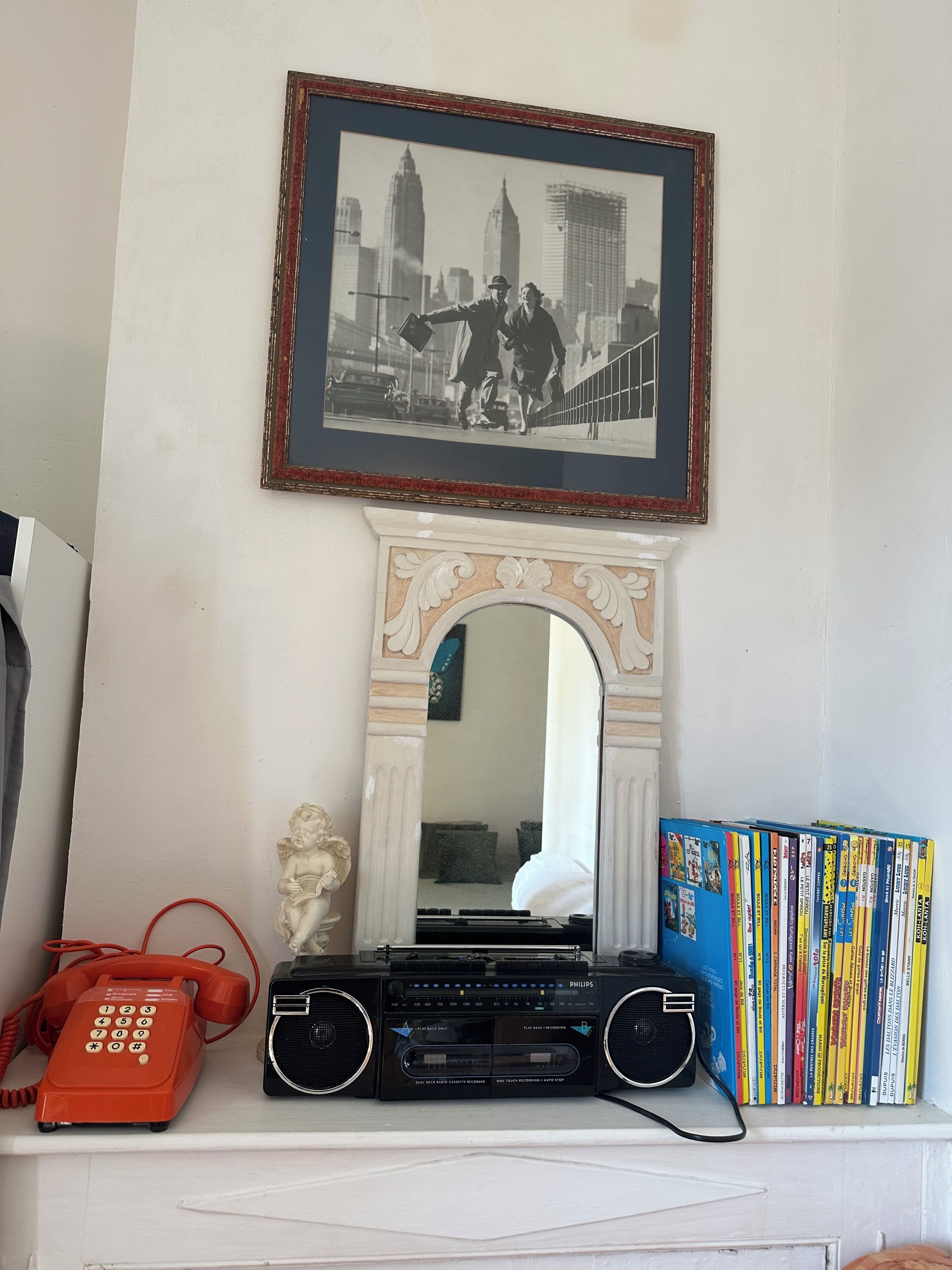 A room with a framed black and white photo, a mirror, a red telephone, a radio, and a bookshelf.