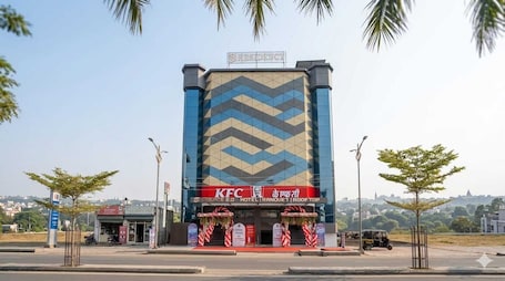 A KFC branded hotel with a modern geometric facade, located at a street corner with palm trees and a clear view of the cityscape in the background.. S S RESIDENCY