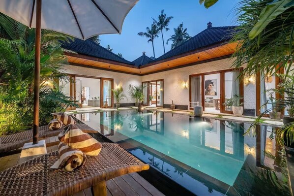 An indoor swimming pool with lounge chairs, surrounded by palm trees and a covered area with wooden beams. - Harmony Villa by DH (Gianyar)