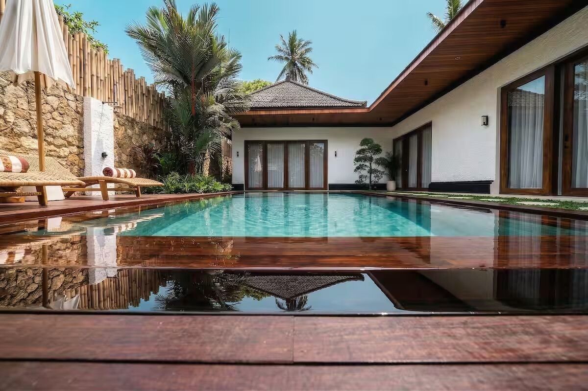 Una piscina con plataforma de madera, pared de piedra y un edificio con grandes ventanales.