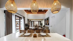 A modern dining area with a central island, three hanging pendant lights, and a view into the dining room. - Harmony Villa by DH (Gianyar)