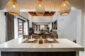 A modern dining area with a central island, three hanging pendant lights, and a view into the dining room. - Harmony Villa by DH (Gianyar)