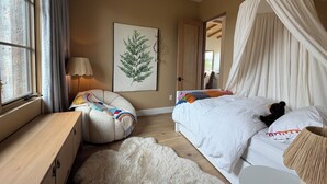 A bedroom with a bed, a bedside table, a lamp, a chair, a rug, a wall-mounted plant, and a door leading to another room. - Sawtooth Sunsets in Stanley, Idaho (Stanley)