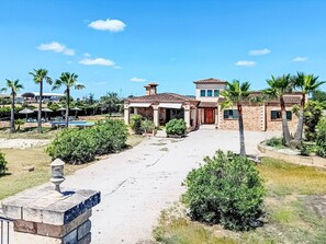 Une grande maison avec une piscine, entourée de palmiers et sous un ciel dégagé.