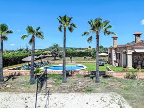 Ein Pool, umgeben von Palmen, ein Steinhaus mit einem Ziegeldach und einem strahlend blauen Himmel.