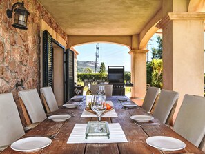 Une table de salle à manger en bois, dressée pour six personnes, avec des chaises, un verre à vin et un barbecue en arrière-plan.
