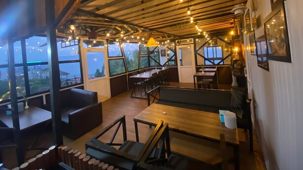 Un restaurante con techo alto, vigas a la vista y luces colgantes. Mesas y sillas dispuestas para comer.
