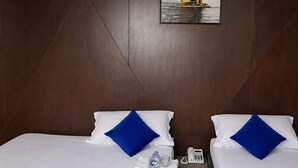 Habitación de hotel con dos camas, cada una con ropa blanca y almohadas azules, una botella de agua, un teléfono y un cuadro con una imagen de un velero en la pared.