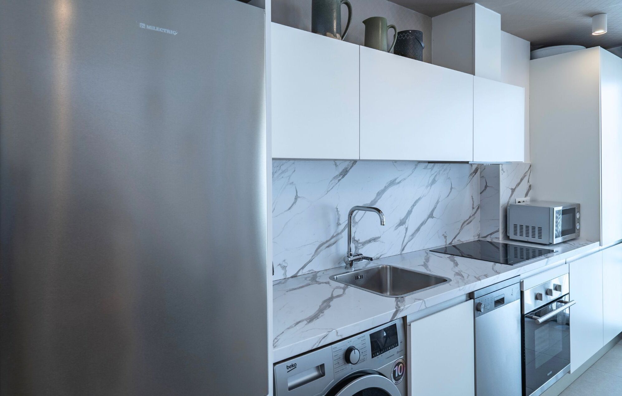 Una cocina moderna con un refrigerador de acero inoxidable, microondas integrado, lavavajillas y un lavabo con salpicadero de mármol.