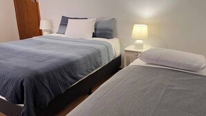Una cama doble con colcha y almohadas azul oscuro, una mesita de noche con lámpara y una puerta de madera al fondo.