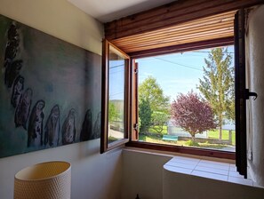Una habitación con un cuadro grande en la pared, una ventana con vista a árboles y una banca afuera, y suelo de baldosas.