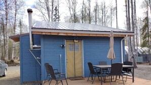 A small cabin with a blue exterior, a wooden deck, a table with chairs, and a covered outdoor seating area.