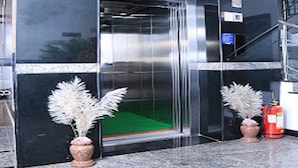 Entrada de un edificio con puerta de ascensor de vidrio, un extintor y dos plantas en macetas.