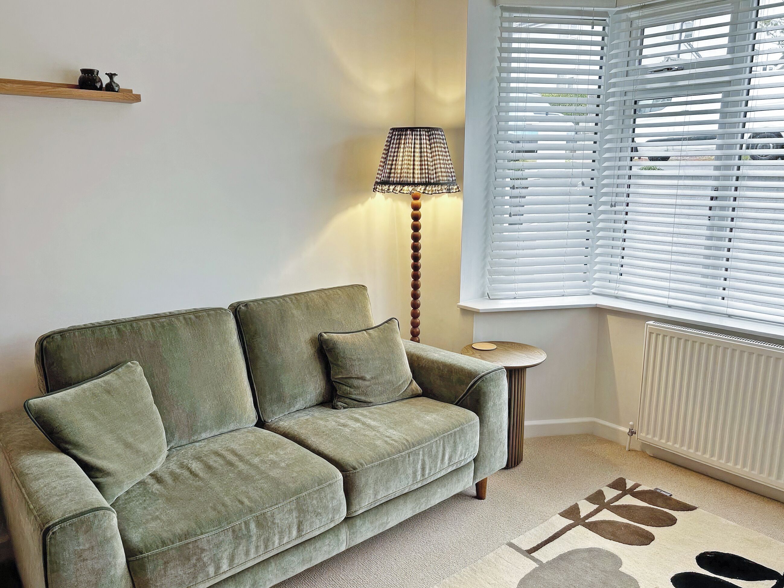 A green sofa with cushions, a small round side table, a floor lamp, and a window with blinds.