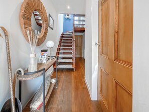 A hallway with a wooden floor, a mirror, a lamp, a desk with a vase and a magazine holder, and a staircase leading to another room. - 3 bedroom accommodation in Stranraer (Stranraer)