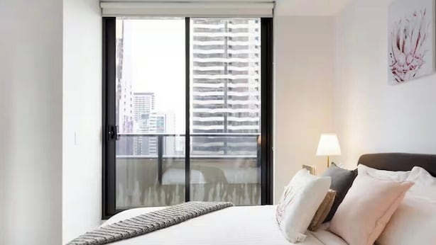 Ein Hotelzimmer mit Bett, Nachttisch, Lampe und Blick auf ein Gebäude durch ein Fenster.