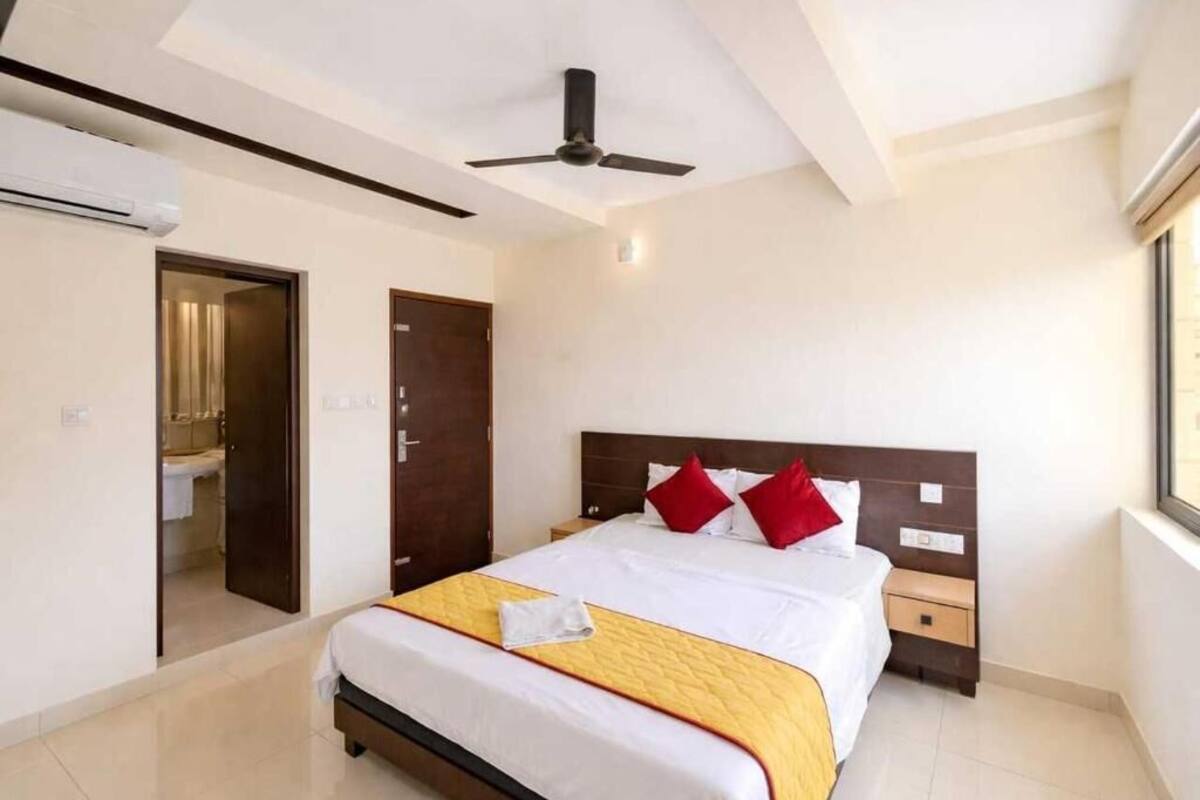 Habitación de hotel con cama, ventilador de techo, ventana y puerta que da a un baño.