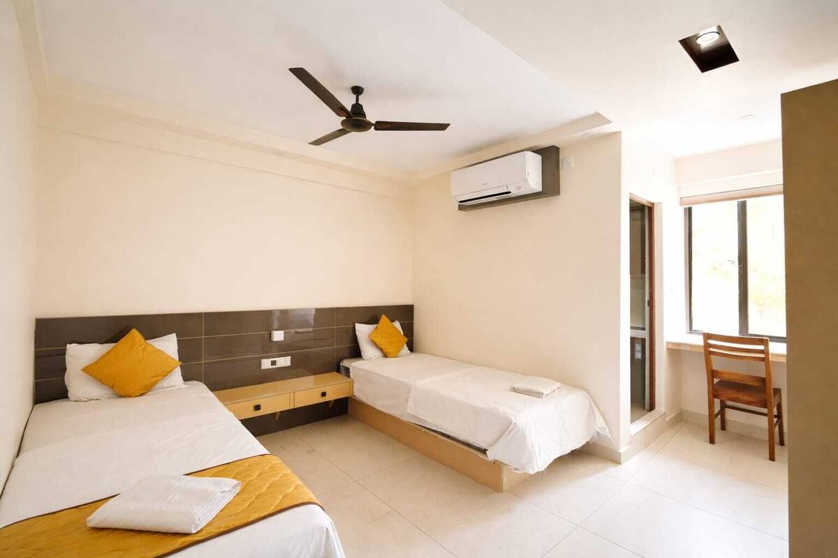 Habitación de hotel con dos camas, un escritorio, una silla, un ventilador de techo, un equipo de aire acondicionado y una ventana.