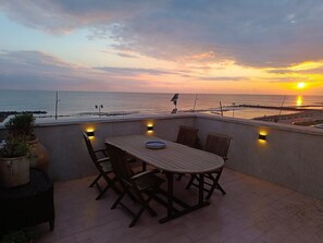 A rooftop terrace with a table and chairs, overlooking the sea at sunset.