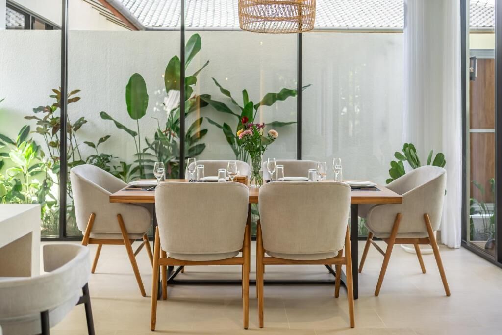 A dining area with a table set for four, surrounded by chairs, with a large window and plants in the background.
